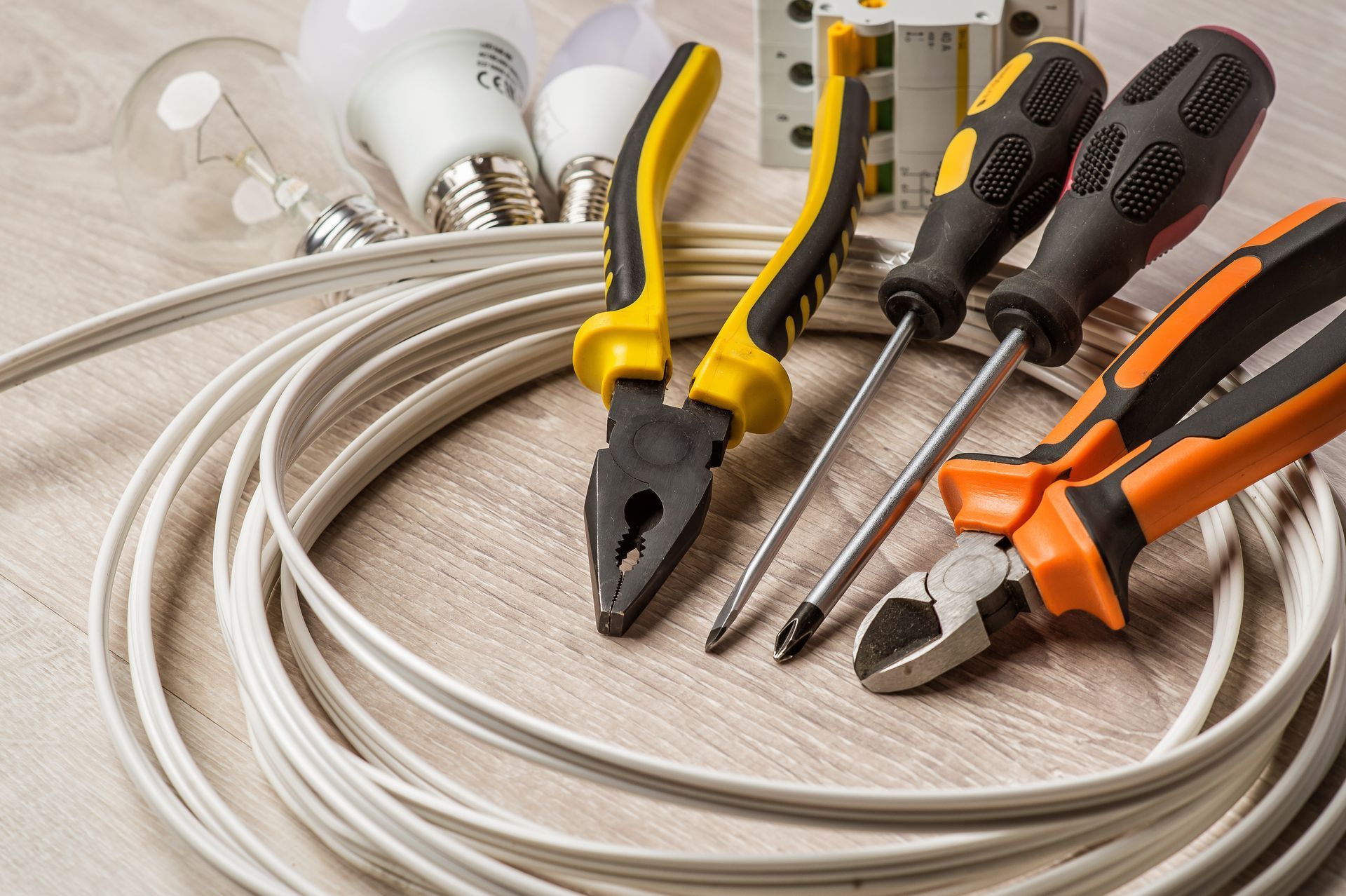 Electrician striping the insulation of wires, working against concrete wall. Coiled white cable with several screwdrivers, pliers, and light bulbs on a wood floor