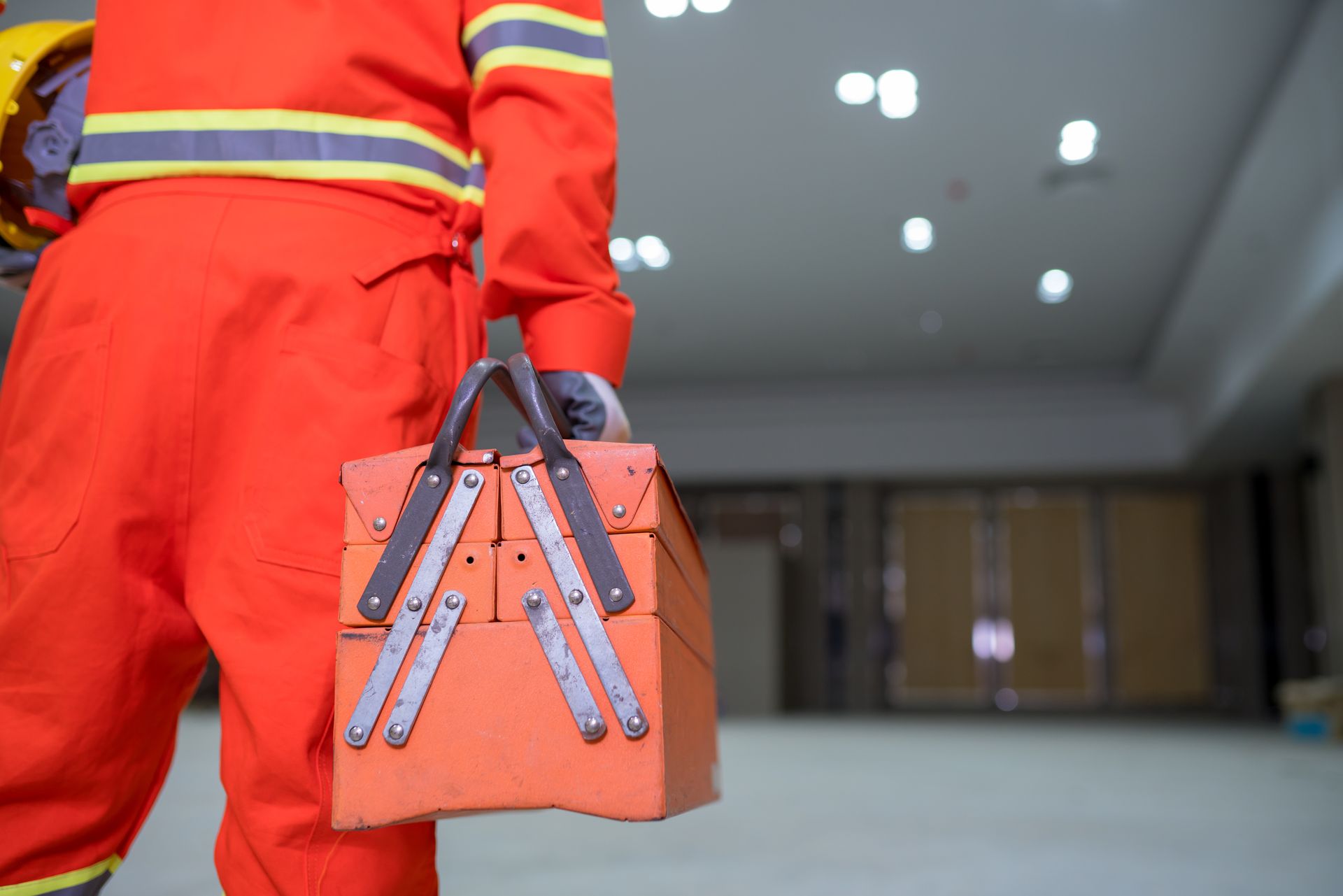 Worker in red safety suit carrying orange toolbox in a building interior.