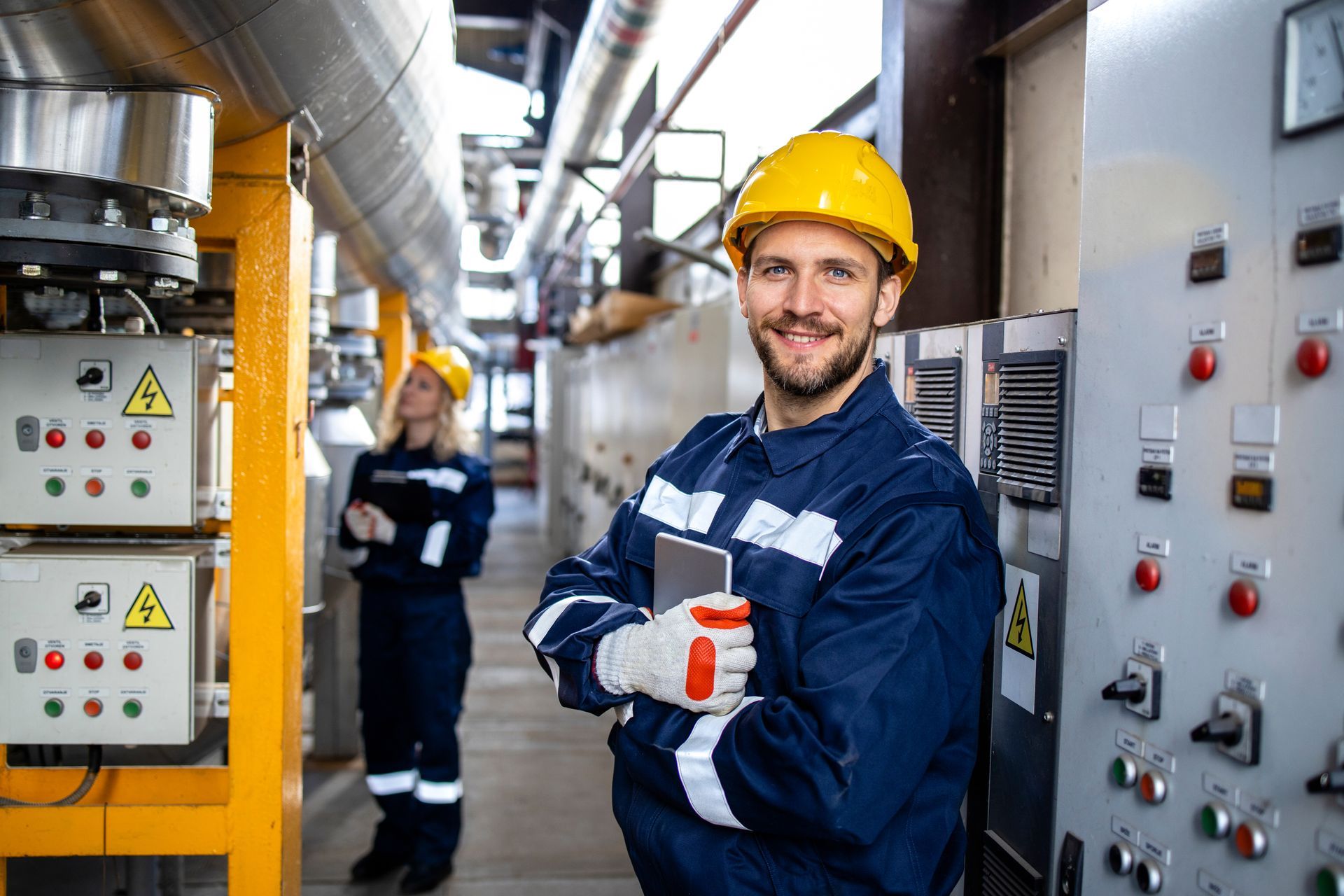 Industrial workers in blue uniforms and yellow hard hats beside control panels in a factory