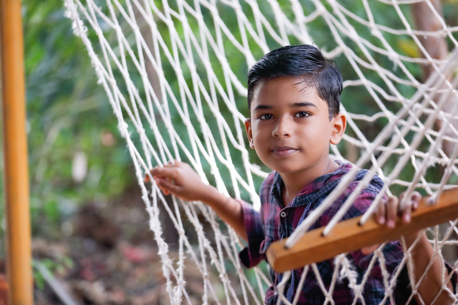 Boy in a hammock, looking at the viewer. He has dark hair and is wearing a plaid shirt.