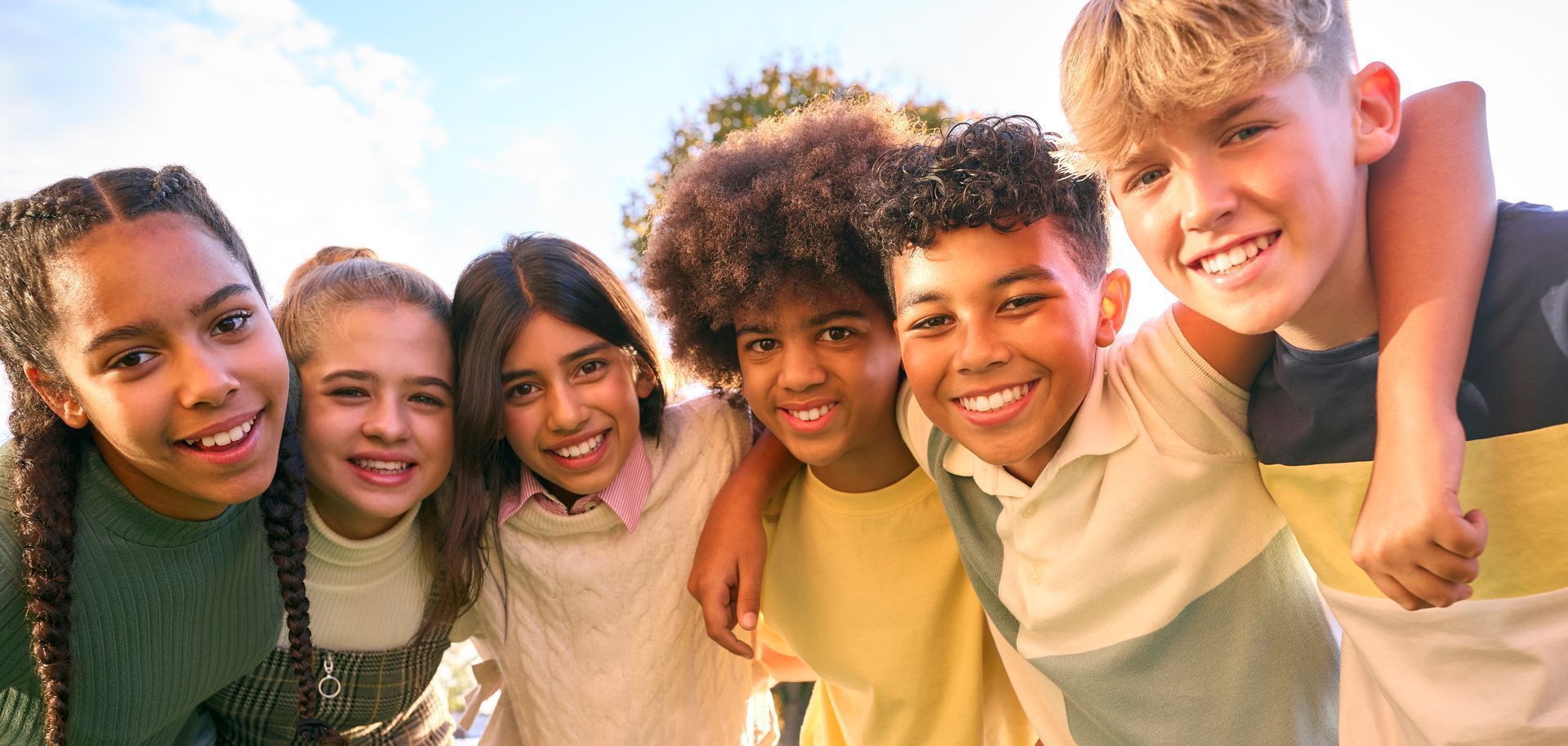 Group of smiling children with arms around each other, outside, under a blue sky.