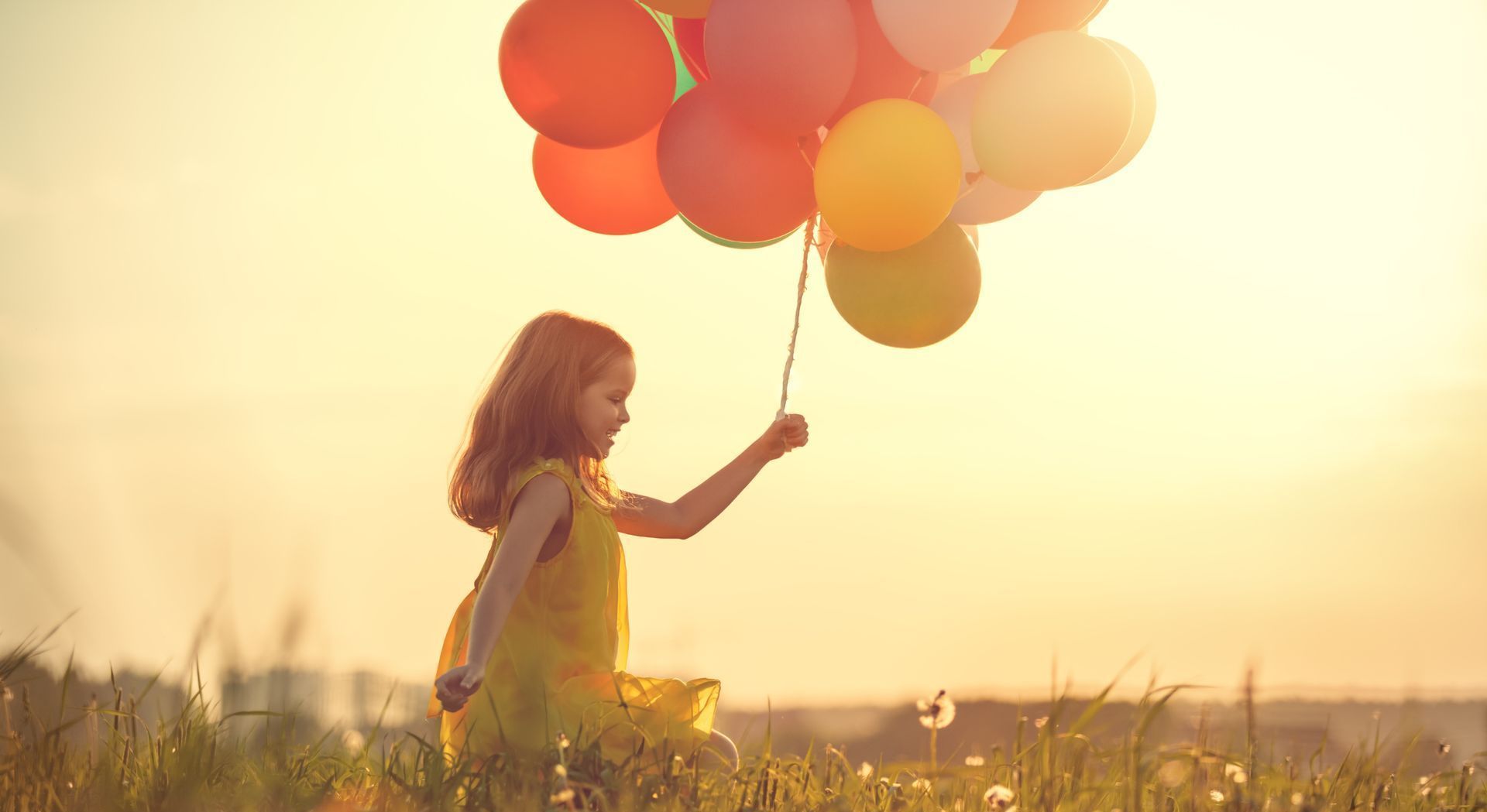 Girl in yellow dress holding colorful balloons in a field, sun setting in the background.