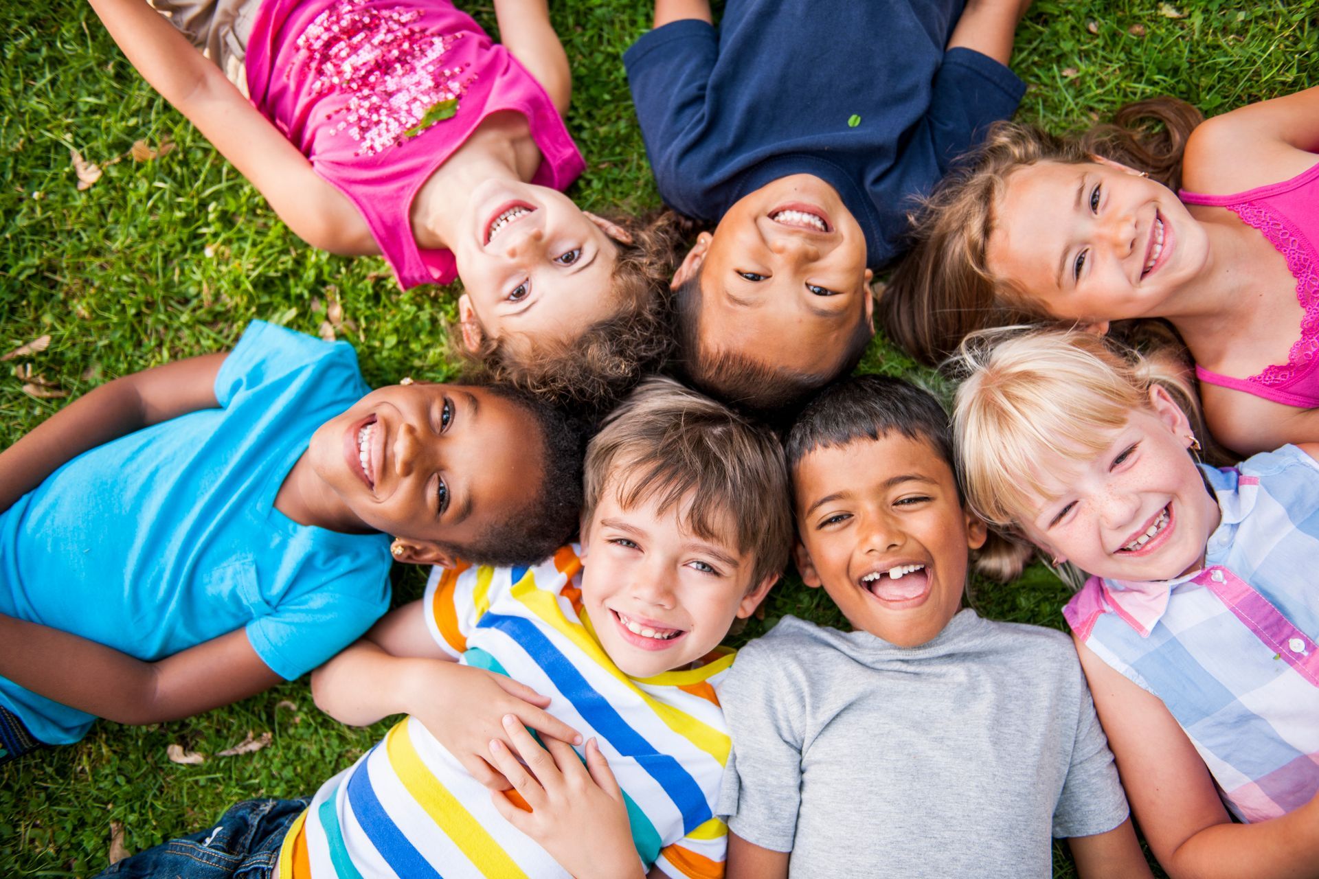 Seven children smiling and lying on grass.