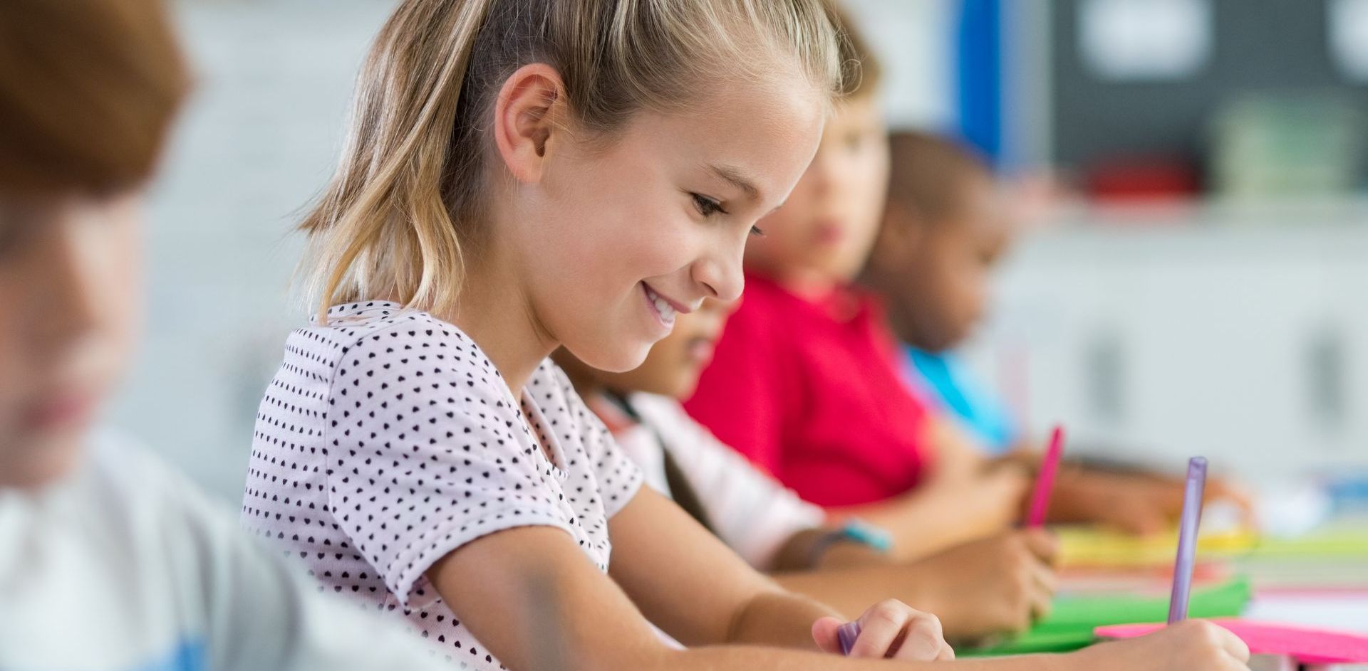 Children writing at their desks in a classroom, the focus on a smiling girl.