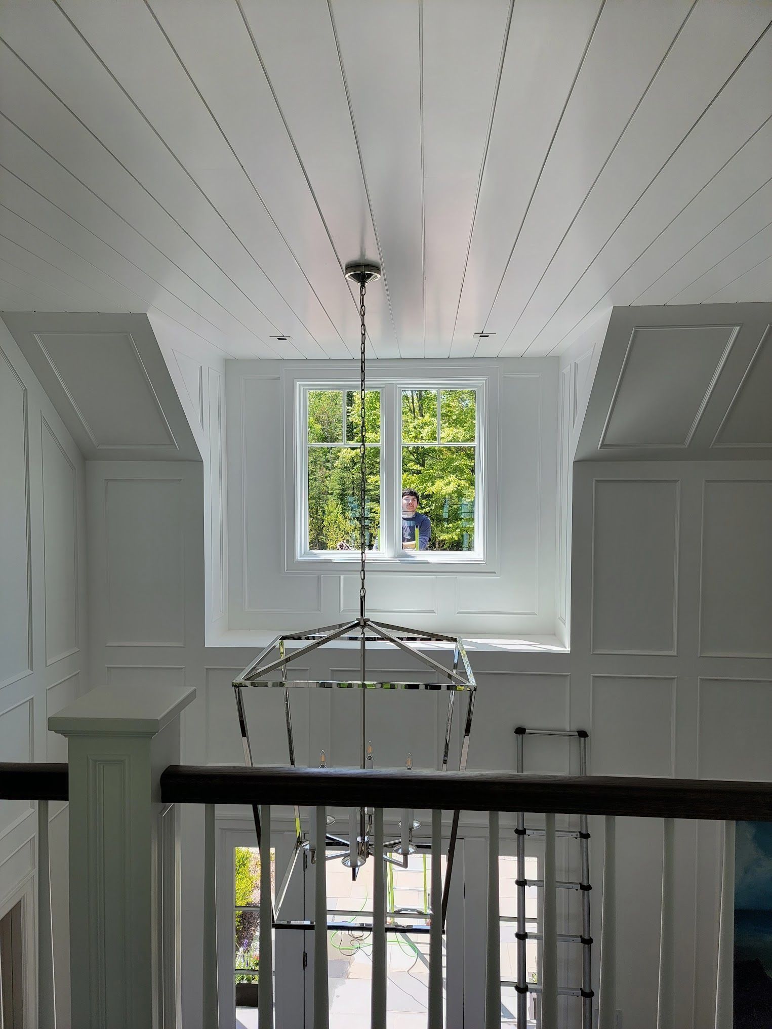 A white hallway with a chandelier hanging in front of a window overlooking trees. The ceiling has wooden planks.