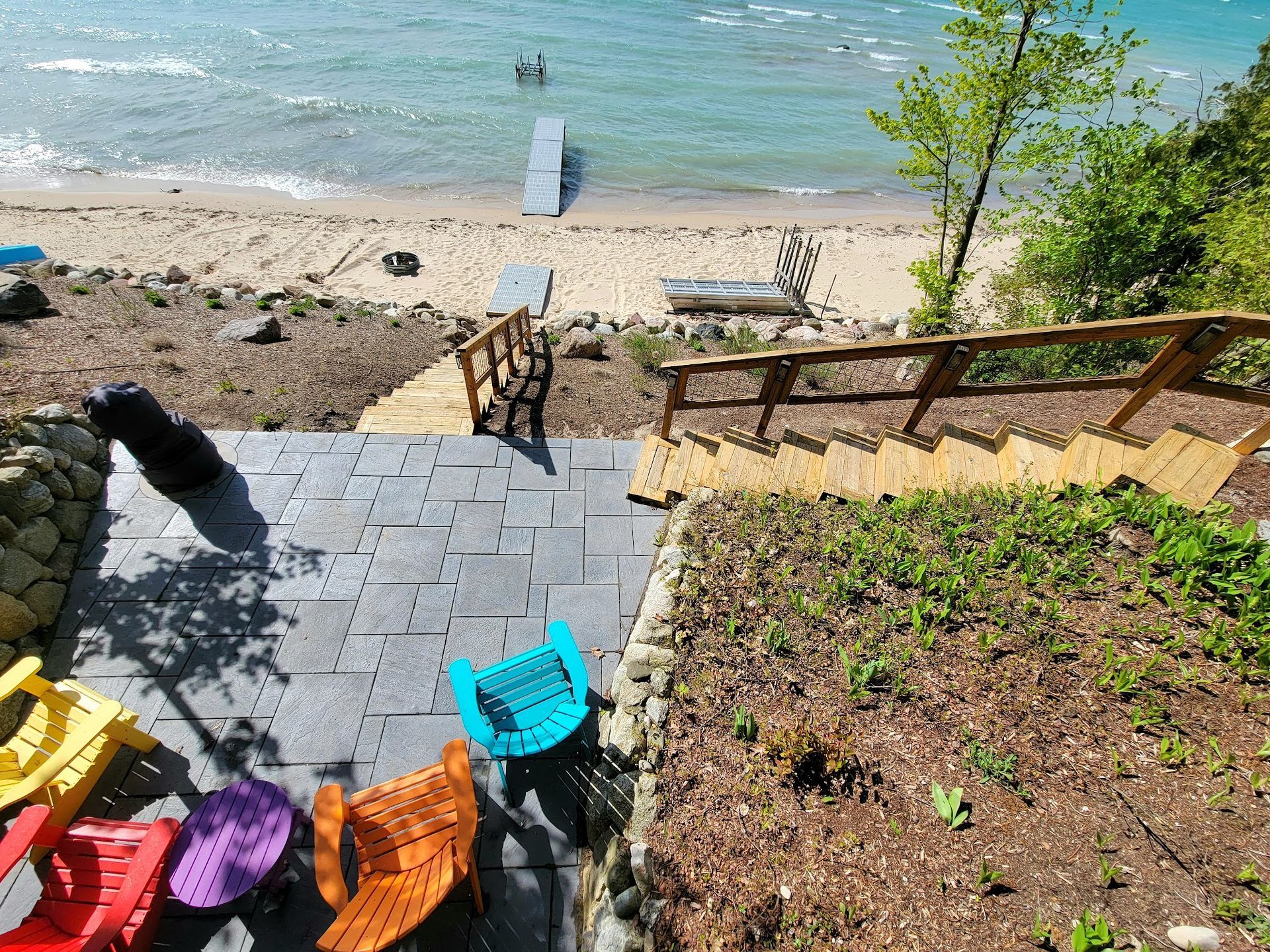 A beach scene with a set of stairs leading down from a patio with colorful chairs. A dock extends into the water.