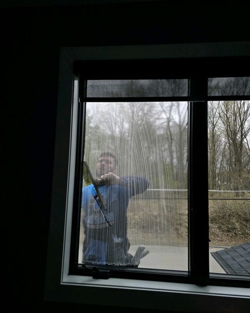 Person cleaning a window with a squeegee. They're visible through the window, in a blue shirt, with a gray backdrop.