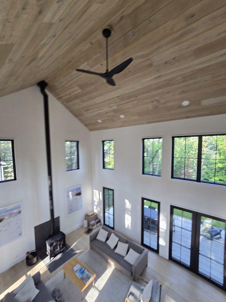 High-ceilinged modern living room with light wood ceiling, black windows, white walls, and a black ceiling fan.