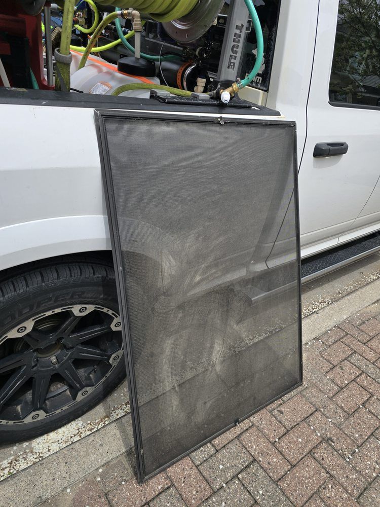 A rectangular black screen leans against a white truck, near a tire on a brick surface. The screen appears dirty.
