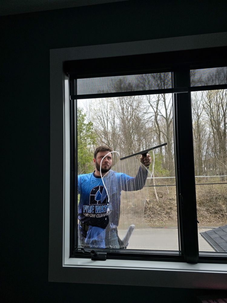 Man cleaning a window with a squeegee. He wears a blue shirt, stands in a room with a window, and has a neutral expression.