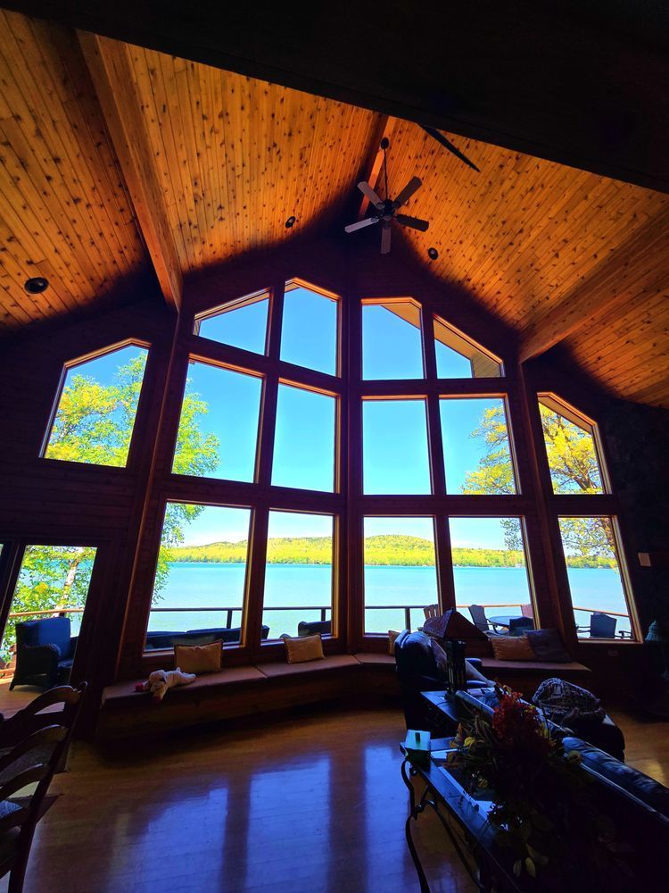 Large window wall of a cabin overlooking a lake. Bright blue sky visible with light filtering through the wooden ceiling.