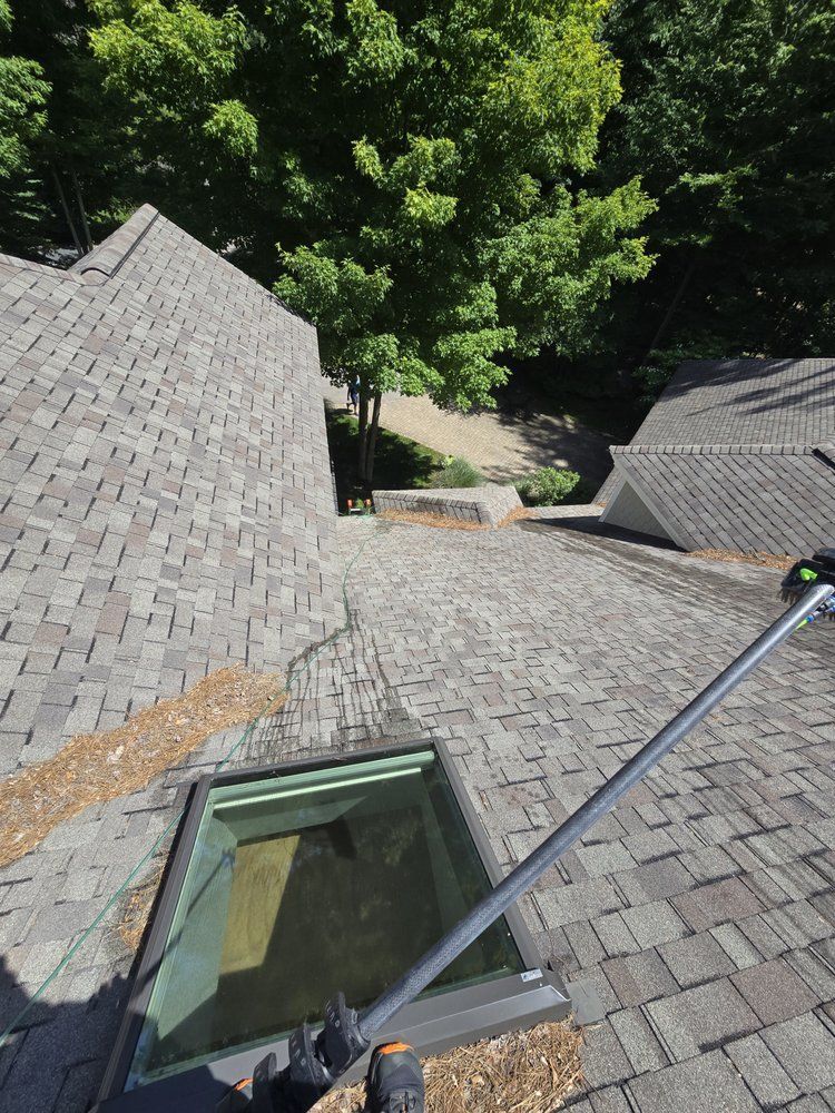 A person on a sloped, shingled roof, cleaning a skylight with a pole. Trees are in the background.