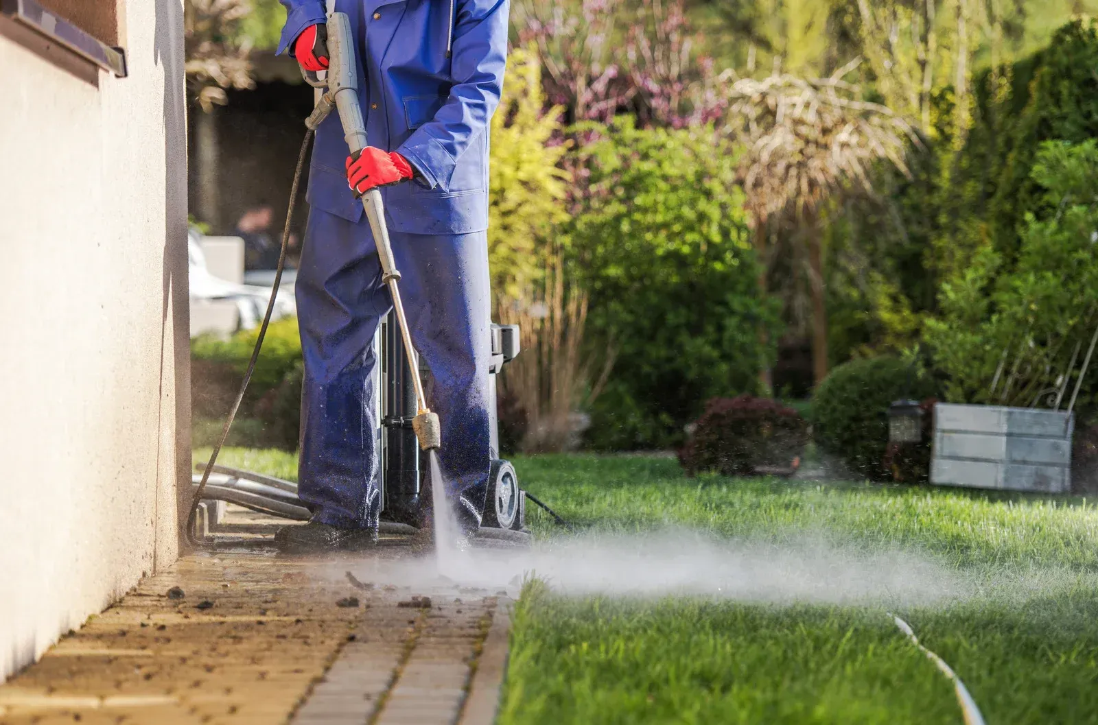 Person in blue overalls pressure washing a brick walkway next to a house with a garden.