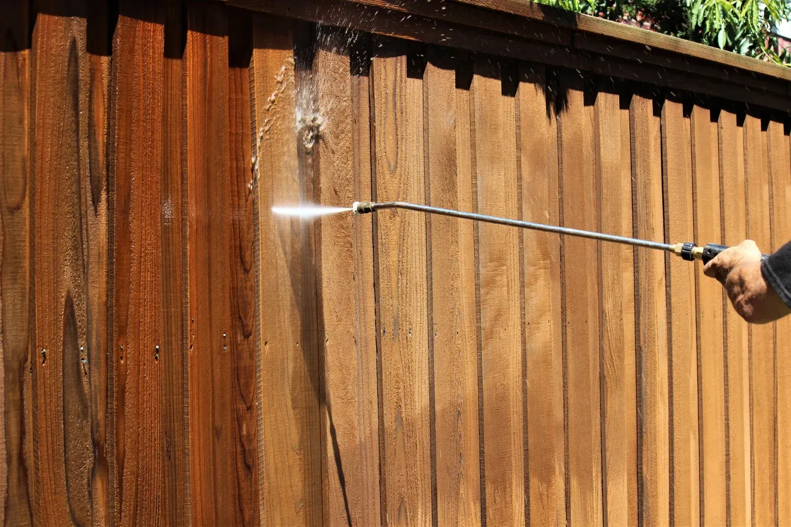 Person using a pressure washer to clean a wooden fence. Water sprays across the stained wood.