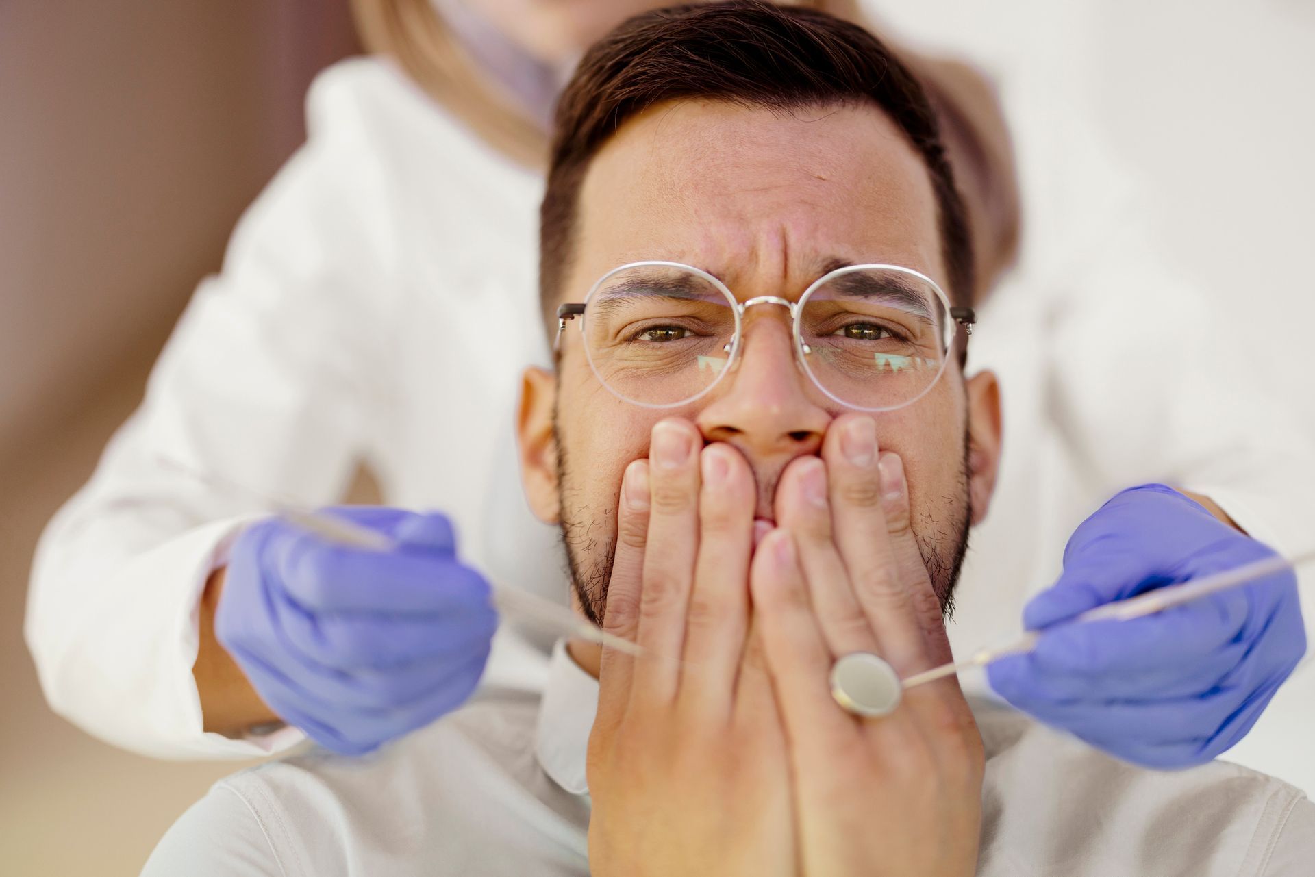 Man with glasses and worried expression covering mouth at the dentist. Dentist in protective gear holds tools.