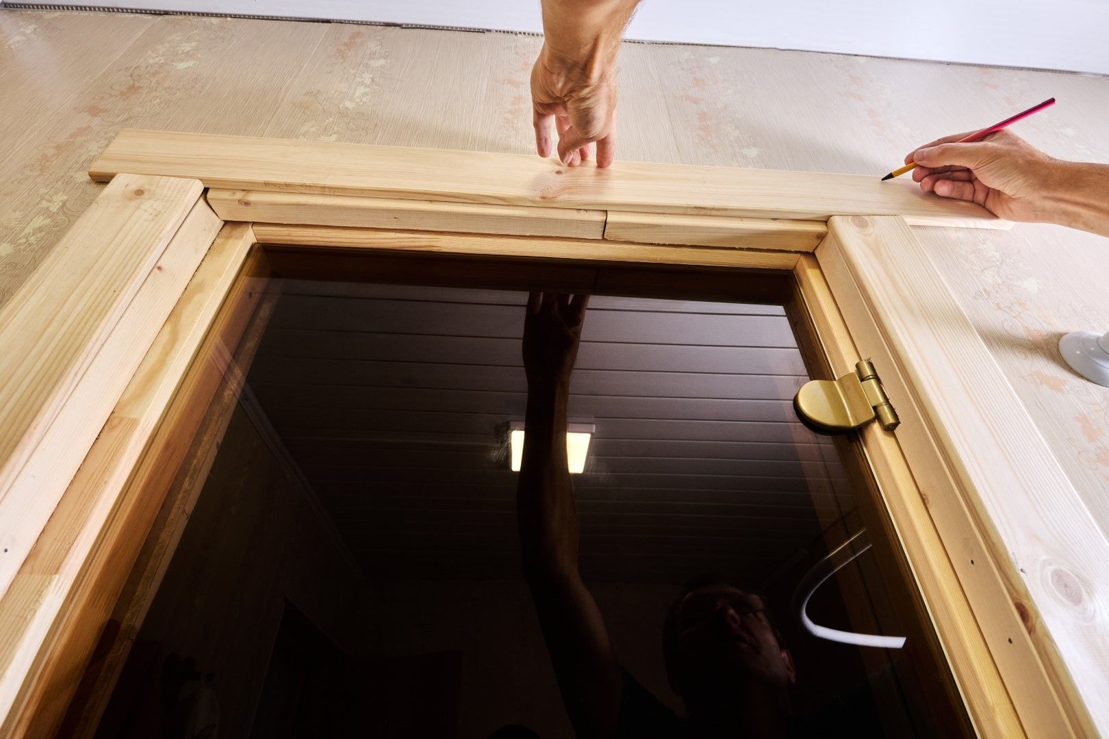 Hands measuring and marking wood trim around a dark-paneled door frame.