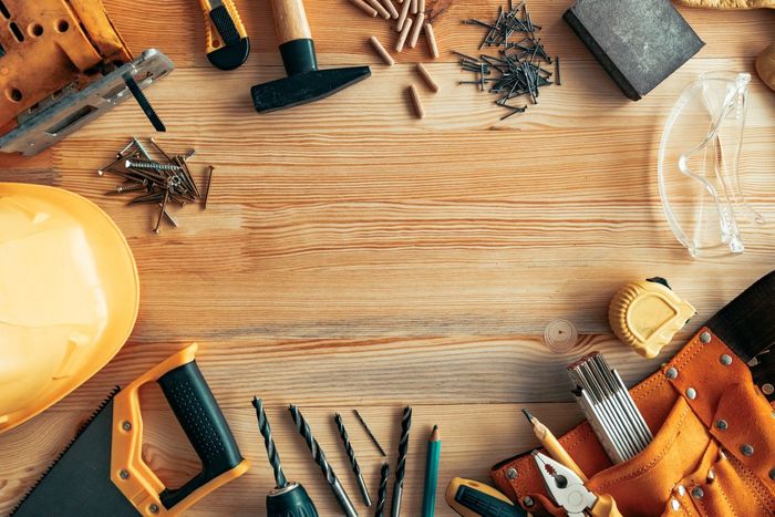 Tools arranged on a wooden surface including a hammer, saw, drill, screws, and a safety helmet.