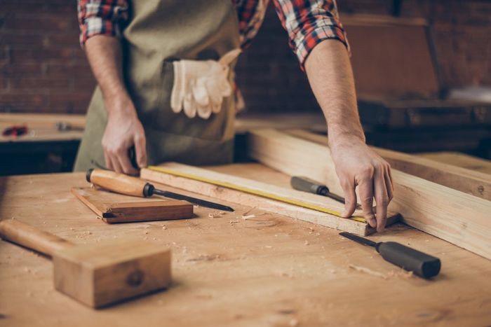Carpenter measuring a wooden plank with a tape measure in a workshop.