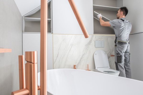 A man installing shelves in a modern bathroom with copper fixtures and a white tub.
