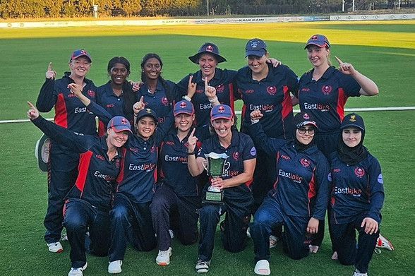 Softball team in navy and red uniforms posing on a field, some kneeling and smiling, with a gold-lit outfield behind them
