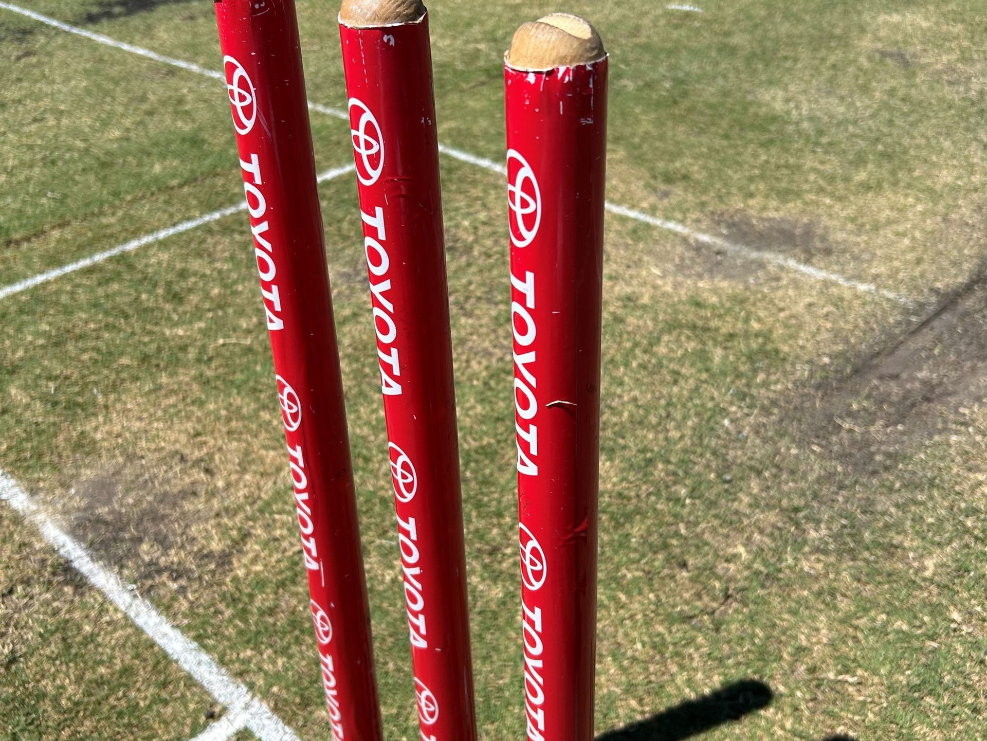 Red cricket stumps with white bails on a grassy field, marked by white boundary lines.
