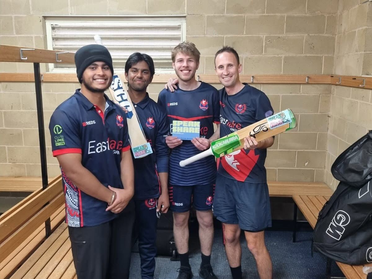 Four teammates in dark jerseys pose indoors, one holding a bright green cricket bat.