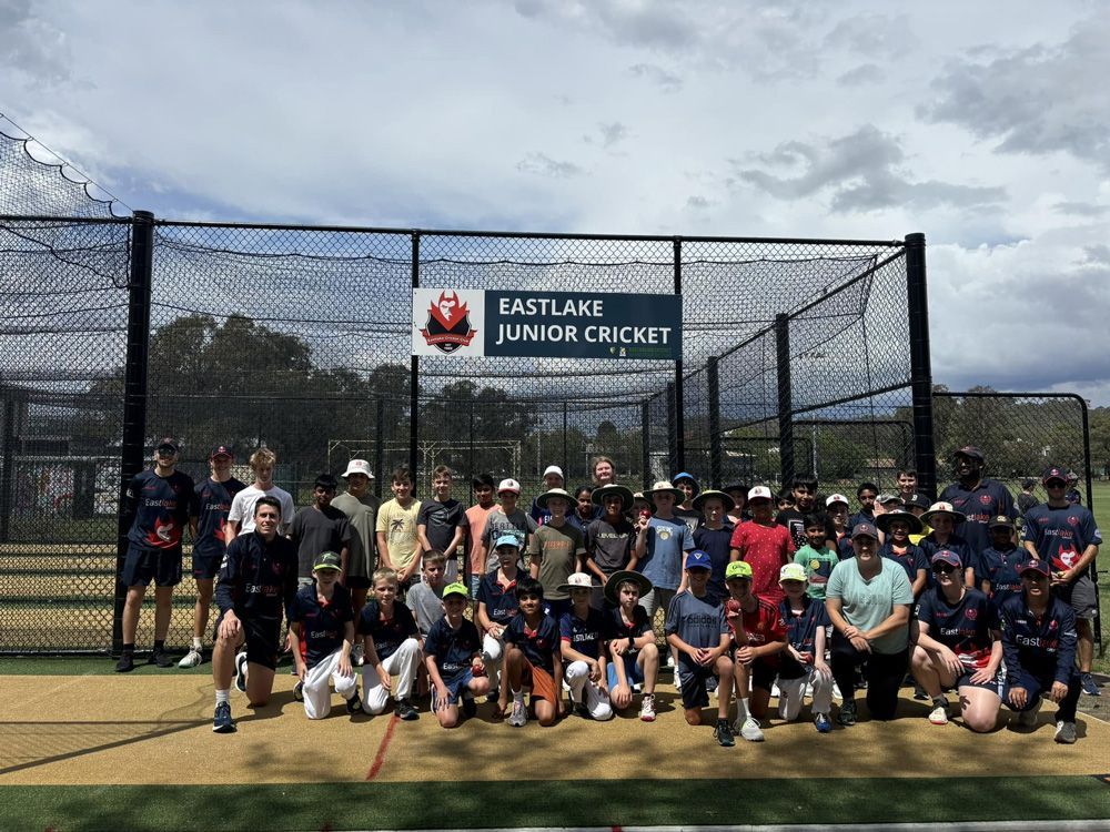 Large group posing on a cricket field in front of a sign reading “Eastlake Junior Cricket”