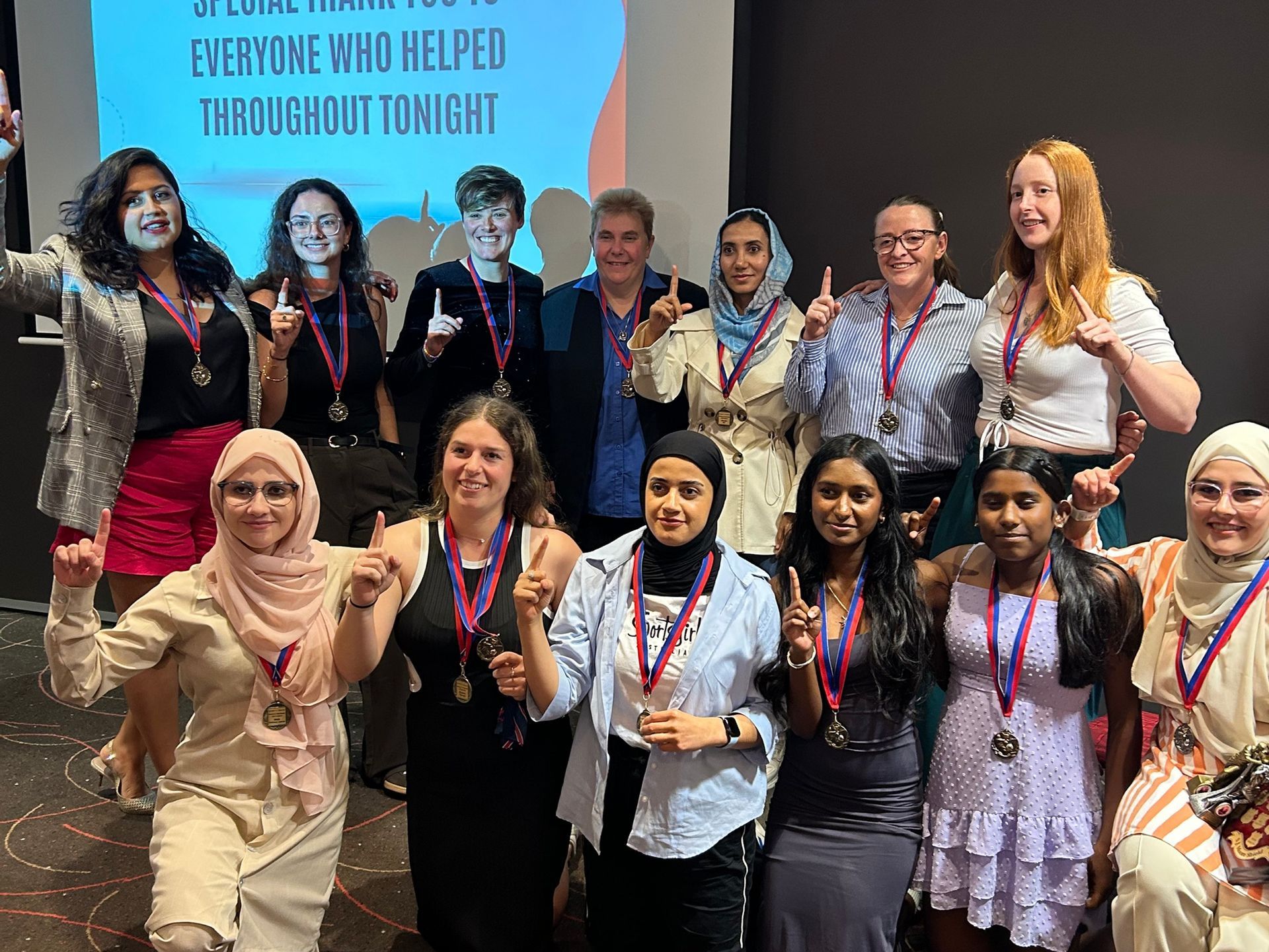 Group of people posing at a community event, smiling with medals in front of a presentation screen