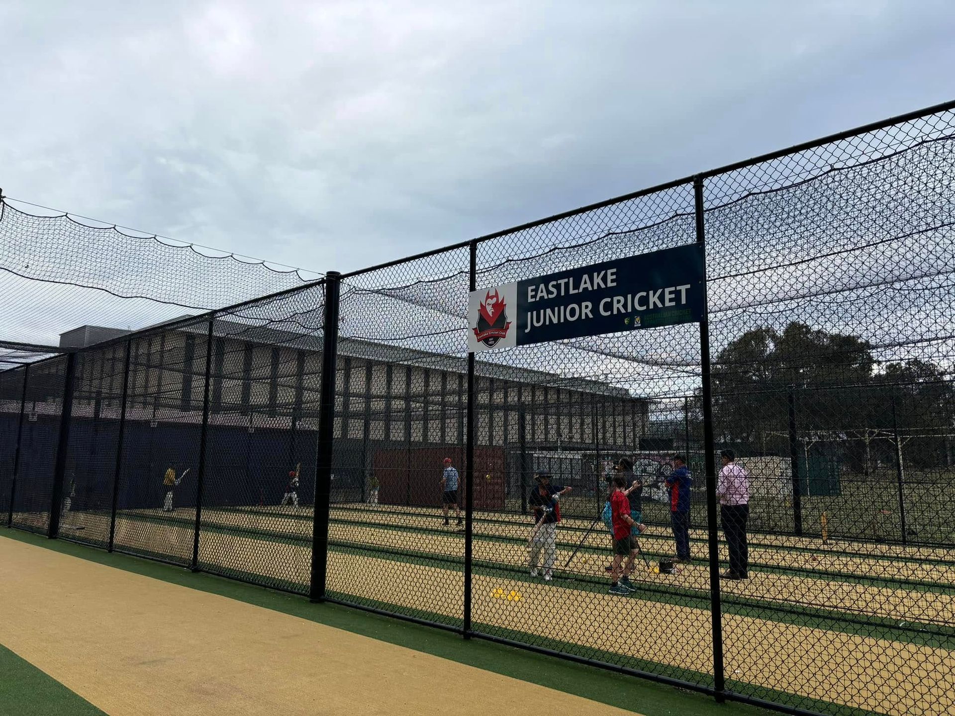 Junior cricket practice nets behind a chain-link fence under a cloudy sky.