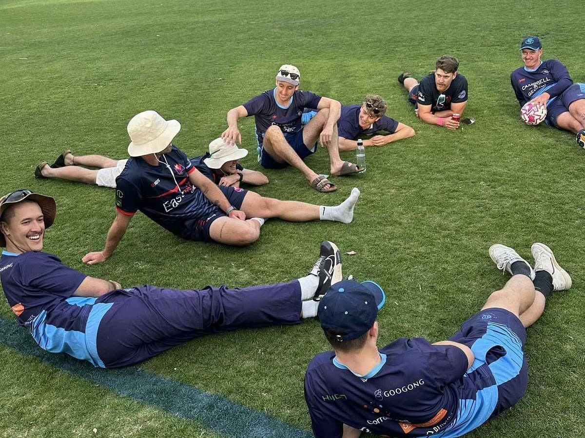 Group of athletes in blue uniforms sitting on a grassy field, relaxing in a circle before practice
