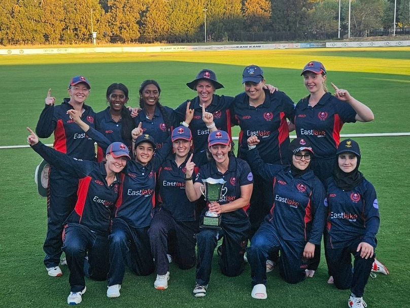 Softball team posing with a trophy on a field, wearing navy uniforms and smiling after a win.