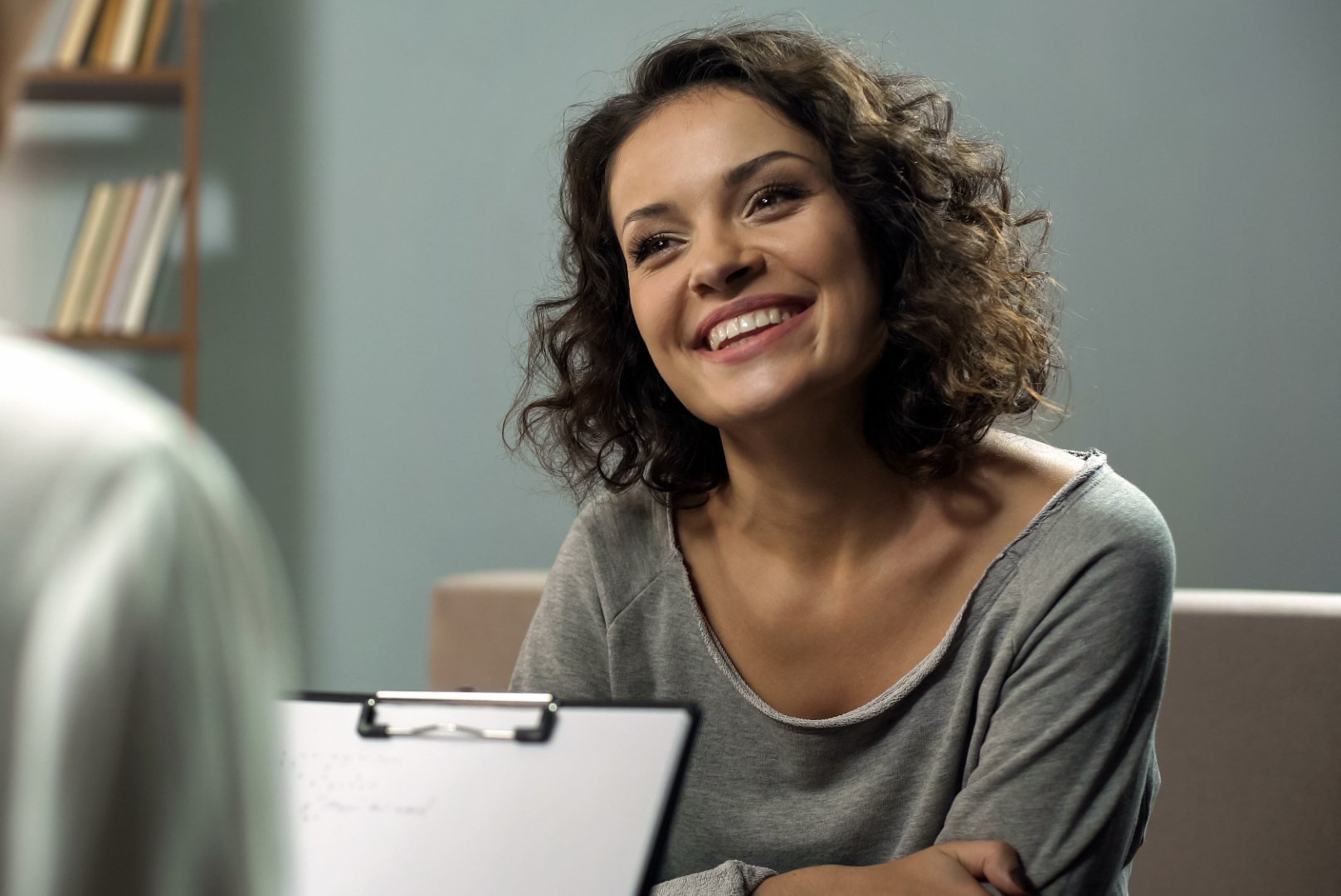 A woman is smiling while sitting on a couch with a clipboard.