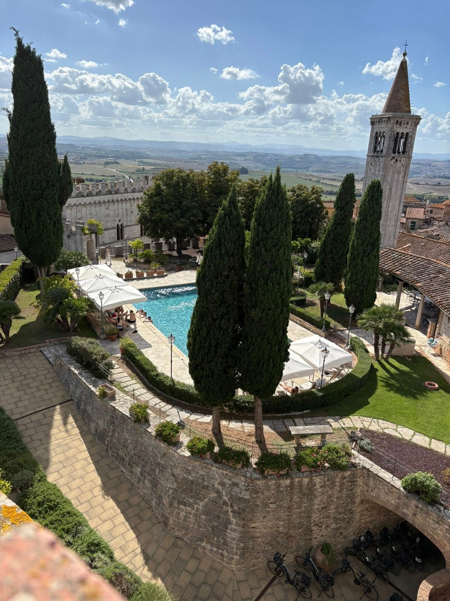 Pool in a walled garden, with tall trees and a distant tower against a sunny sky.