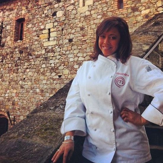 Woman in chef's coat, standing in front of a stone wall. She is smiling with one hand on her hip.