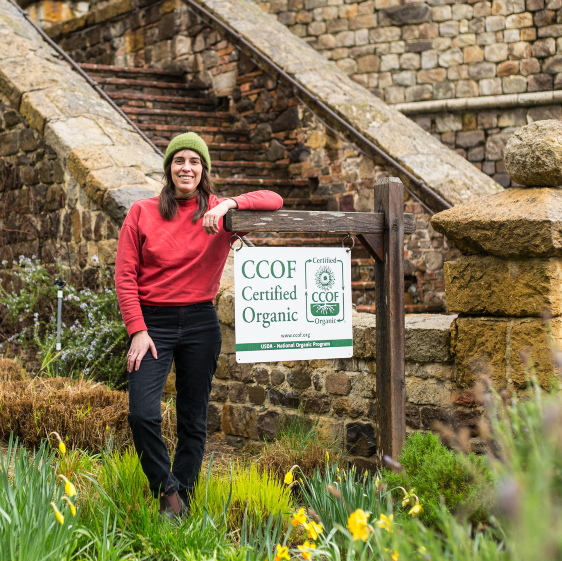 Woman in red sweater and green hat beside a CCOF Certified Organic sign near stone steps.