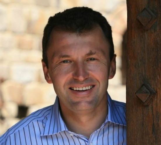Man smiling, leaning on a wooden doorframe, blue striped shirt, brick background.
