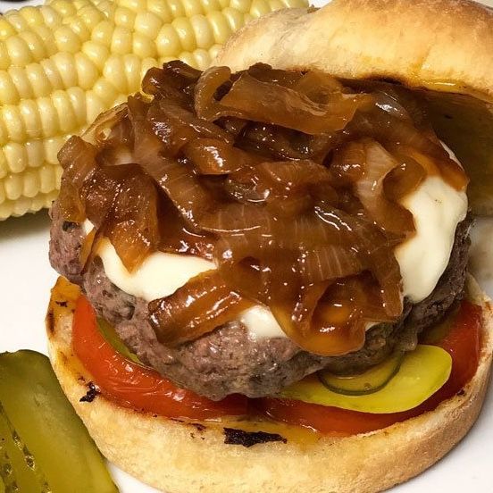 Burger with caramelized onions, cheese, tomato, and pickle on a bun, served with corn on the cob.