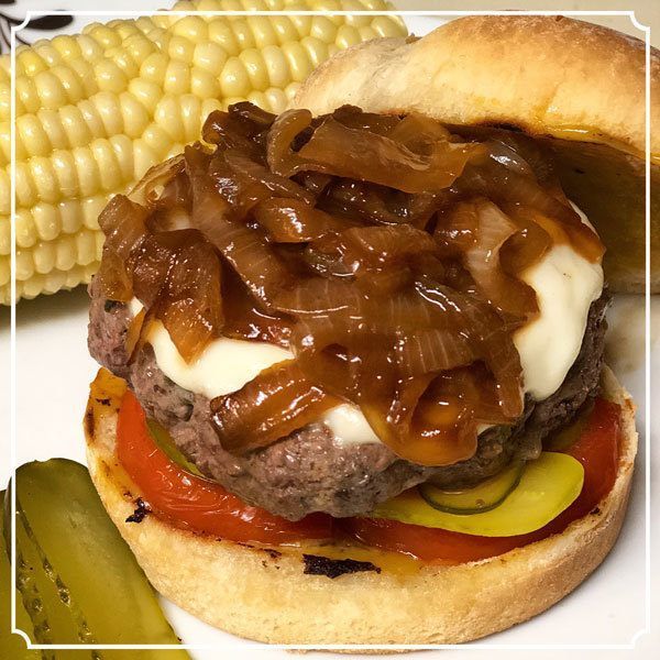 Burger with caramelized onions, cheese, tomato, and pickle, served with corn on the cob.