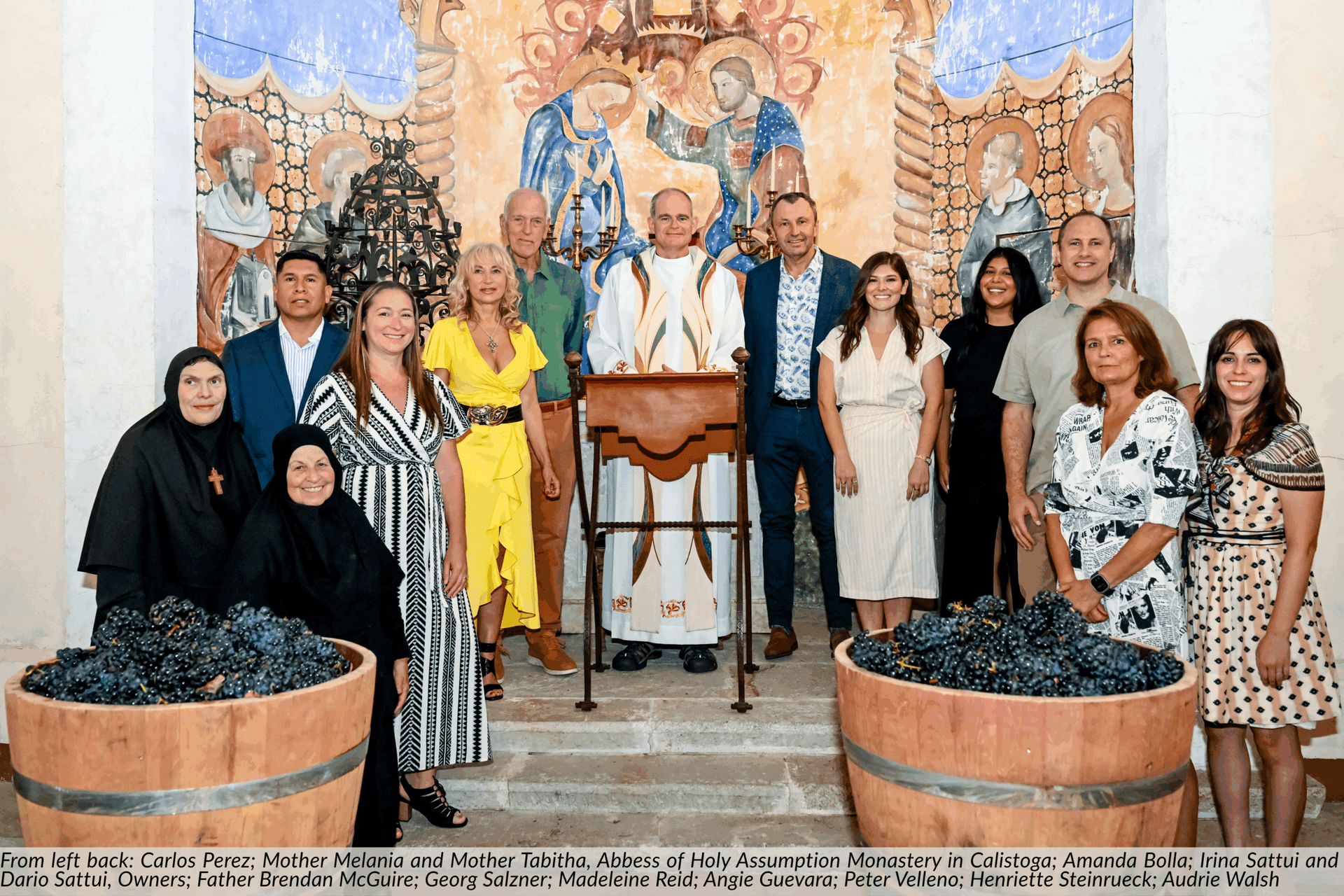 Group of people by barrels of grapes, with a religious figure standing at a podium in front of a mural.