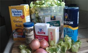 Ingredients for a recipe: chicken stock, lettuce, salt, shallots, butter, black pepper, and herbs on a cutting board.