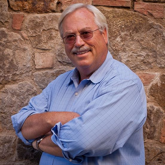 Man with mustache wearing glasses, blue shirt, arms crossed, smiling in front of a stone wall.