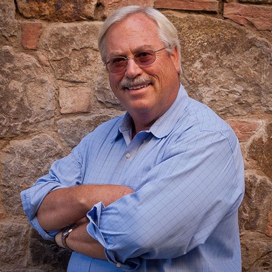 Man with glasses and mustache, arms crossed, smiling, wearing a blue shirt, leaning against a stone wall.