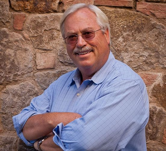 Man with glasses, arms crossed, smiling. Wearing a light blue button-down shirt, standing in front of a stone wall.