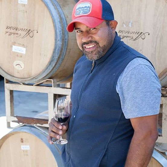 Man holding wine glass smiles, standing near wine barrels, wearing a vest and cap.
