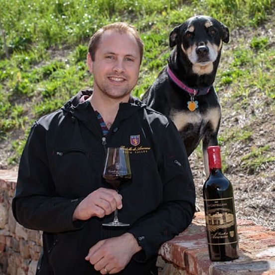 Man holding wine glass with dog and bottle of wine on a brick wall, outdoors.