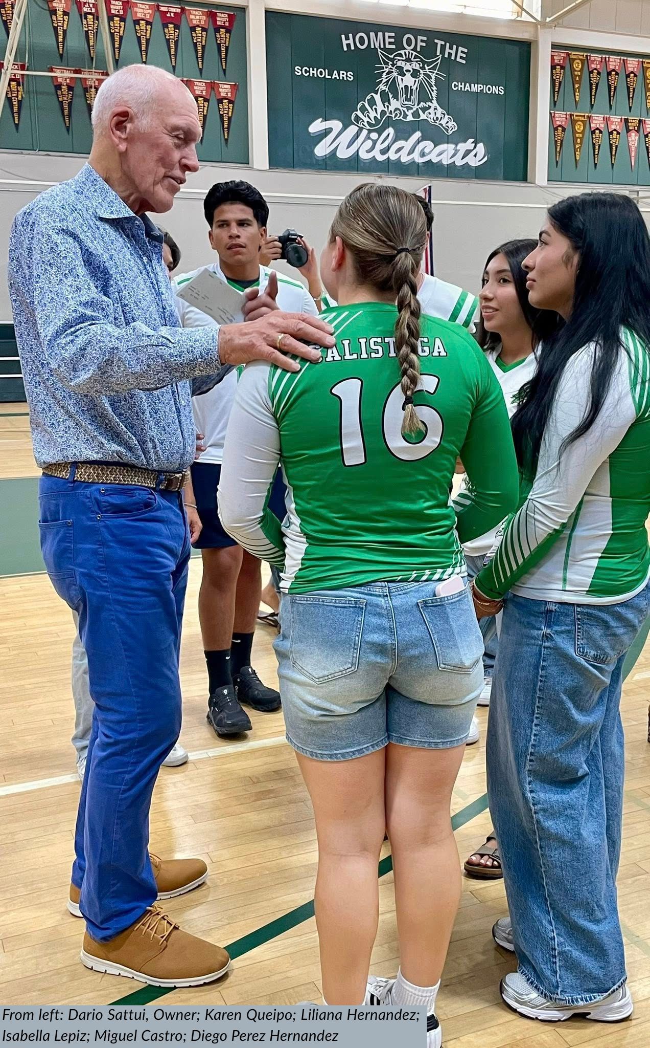 Man talking to a group of people wearing green and white jerseys in a gymnasium.