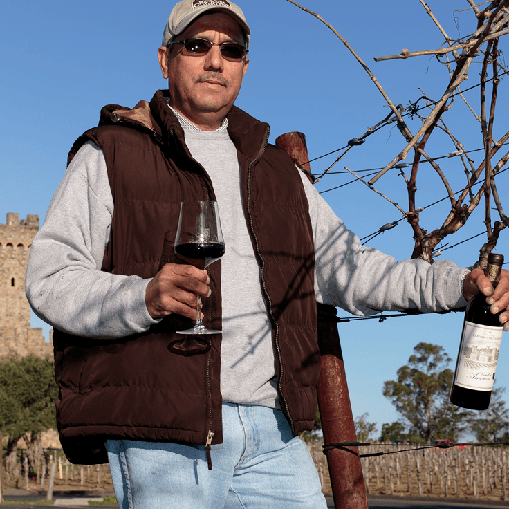 Man holding wine glass and bottle at vineyard. Brown vest, light blue jeans, and sunglasses.