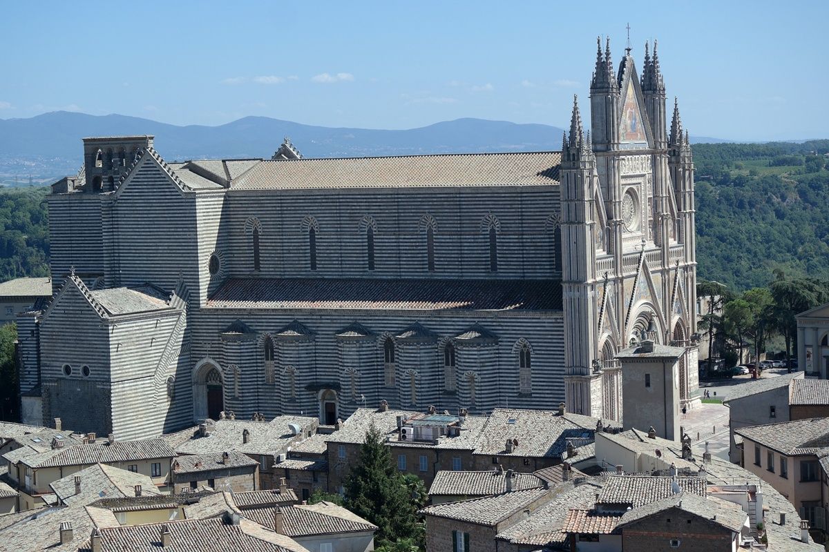 Orvieto Cathedral, Italy. Striking facade with intricate detail, striped side, overlooking rooftops, against a blue sky.