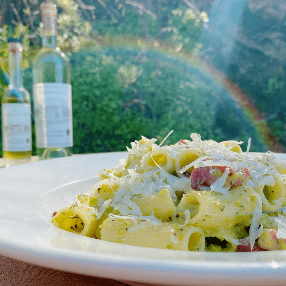 Pasta dish with cheese, bacon, and herbs, with wine bottles and rainbow in background.