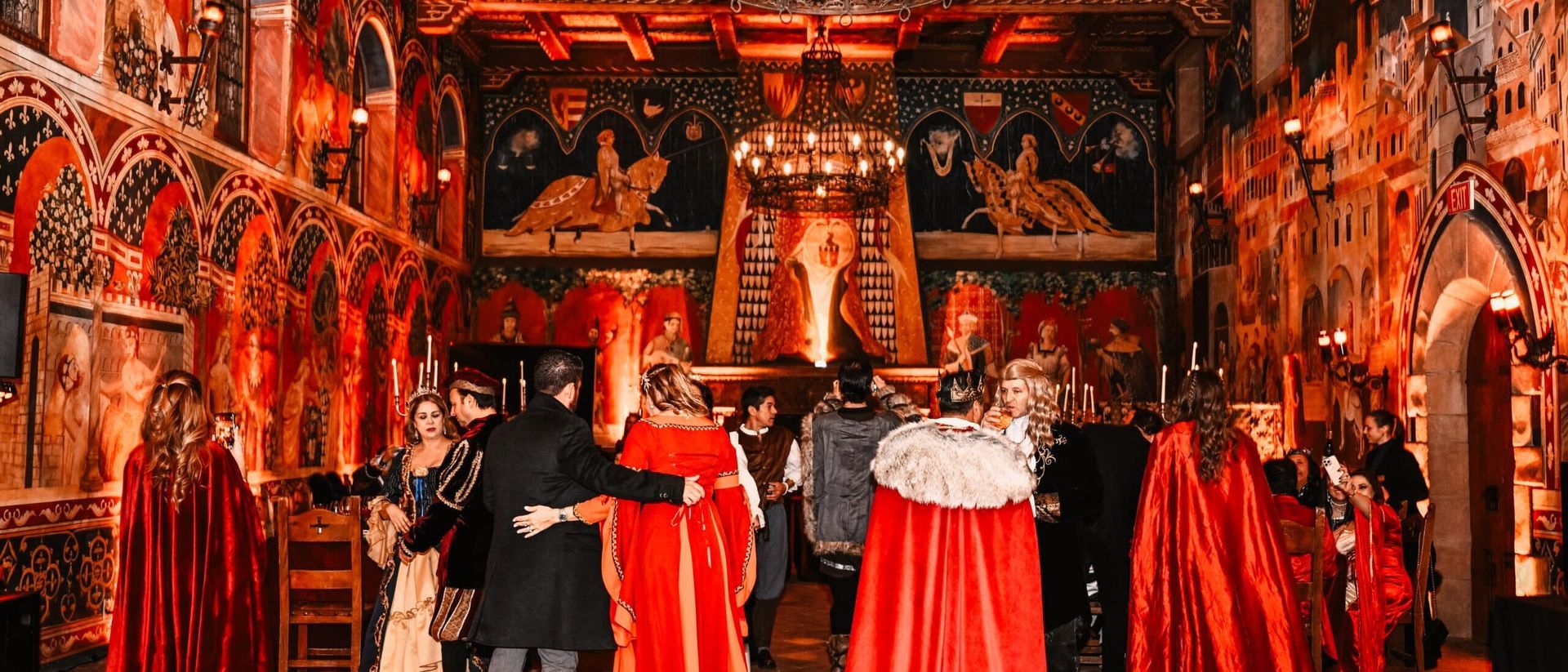 People in red cloaks gather in a gothic hall with elaborate wall art and a large chandelier.